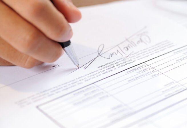 Closeup of person signing document with ball pen. Contract lying on table. Agreement concept. Cropped view.