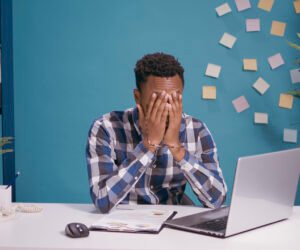 Frustrated worker feeling exhausted about office work, using laptop computer at desk. Tired employee working on business plan, feeling angry and disappointed about financial mistake.